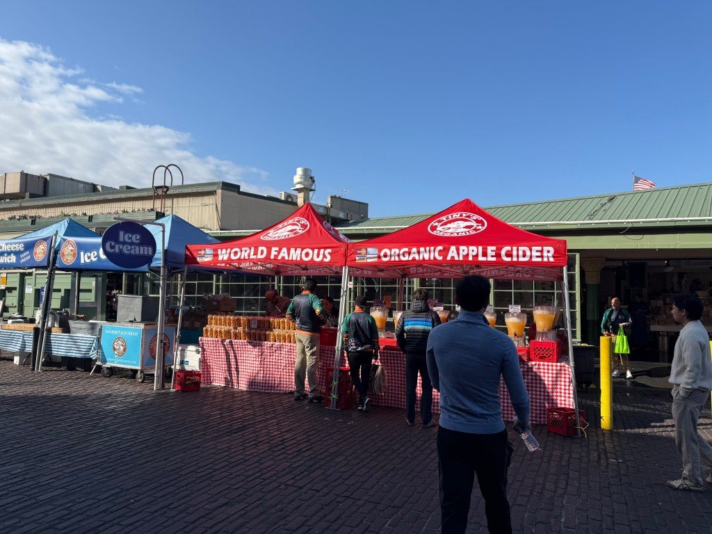 Stalls at Pike Place Market featuring tents for ice cream, organic apple cider, and other local food offerings, with visitors enjoying the vibrant atmosphere.