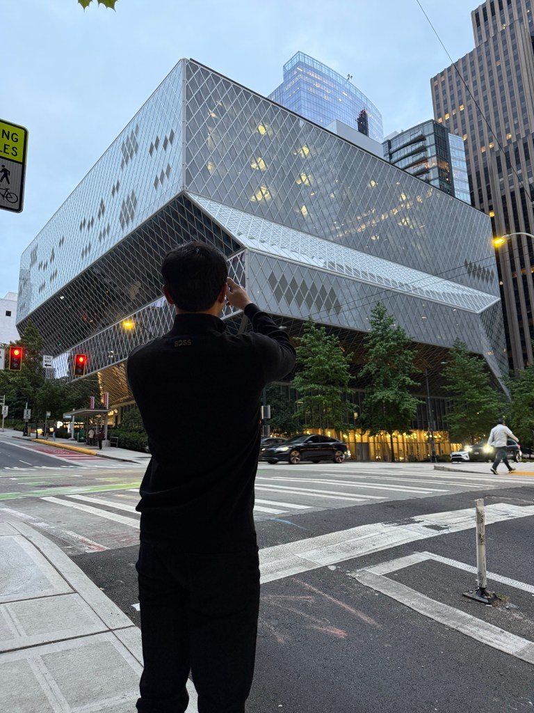 A person standing on the street photographing the Seattle Central Library, showcasing its modern architectural design with a diamond-patterned glass exterior.
