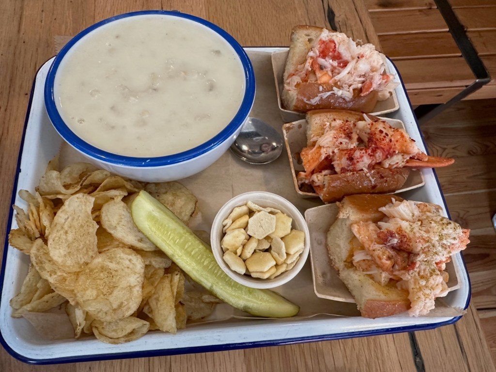 A tray featuring New England clam chowder in a blue-rimmed bowl, two lobster rolls topped with large pieces of lobster meat, a side of potato chips, a pickle, and a small dish of crackers.