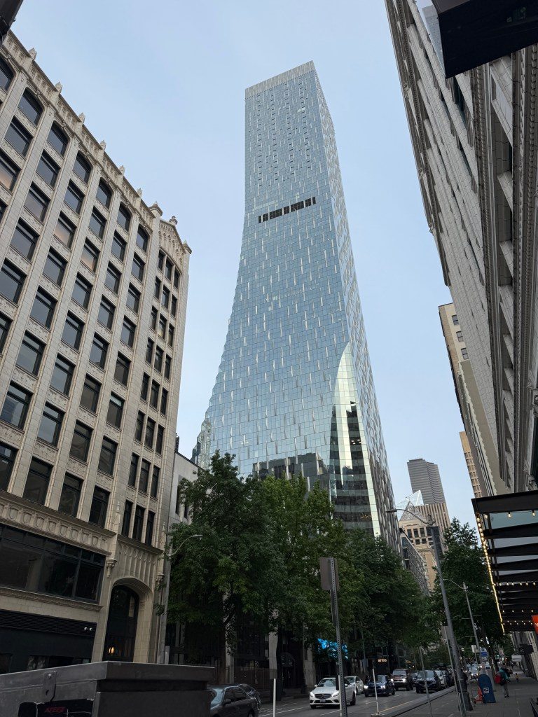 A modern glass skyscraper in Seattle, towering above surrounding older buildings, with trees lining the street below.