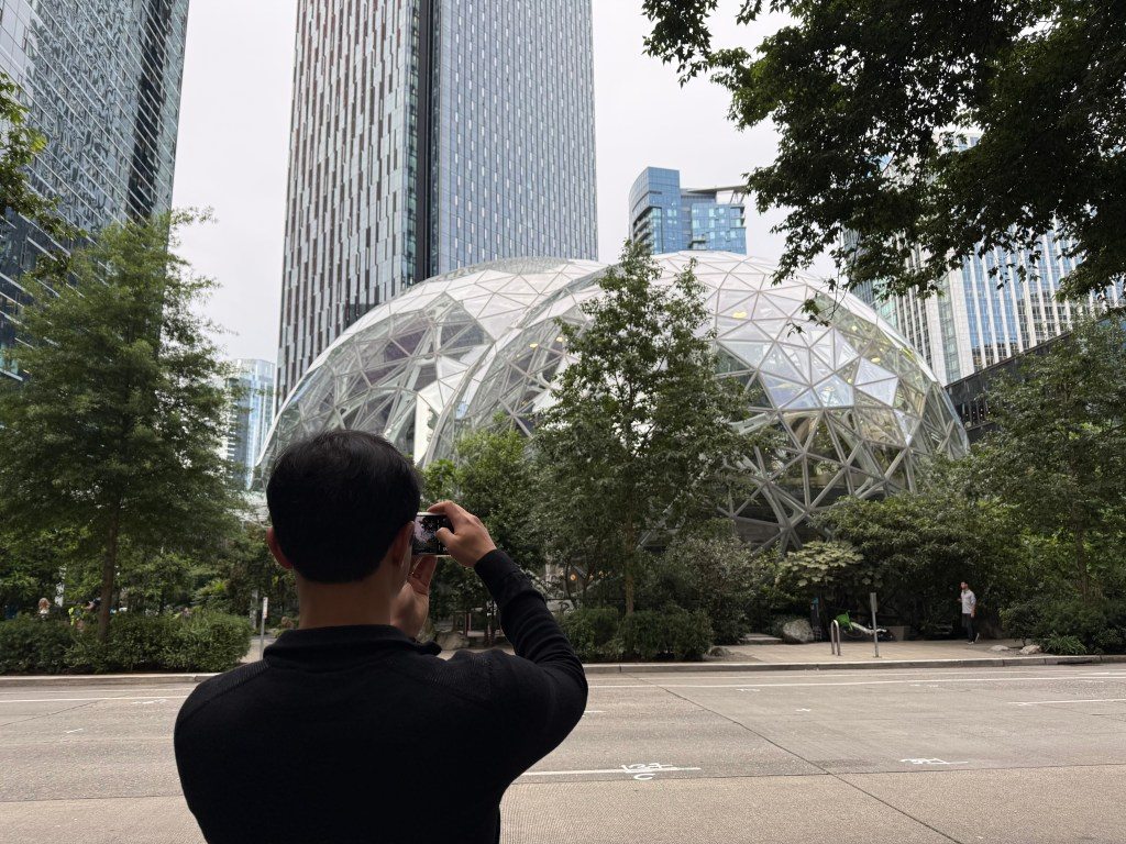 A person taking a photo in front of the Amazon Spheres, a distinctive glass and steel structure surrounded by lush greenery and modern buildings in Seattle.
