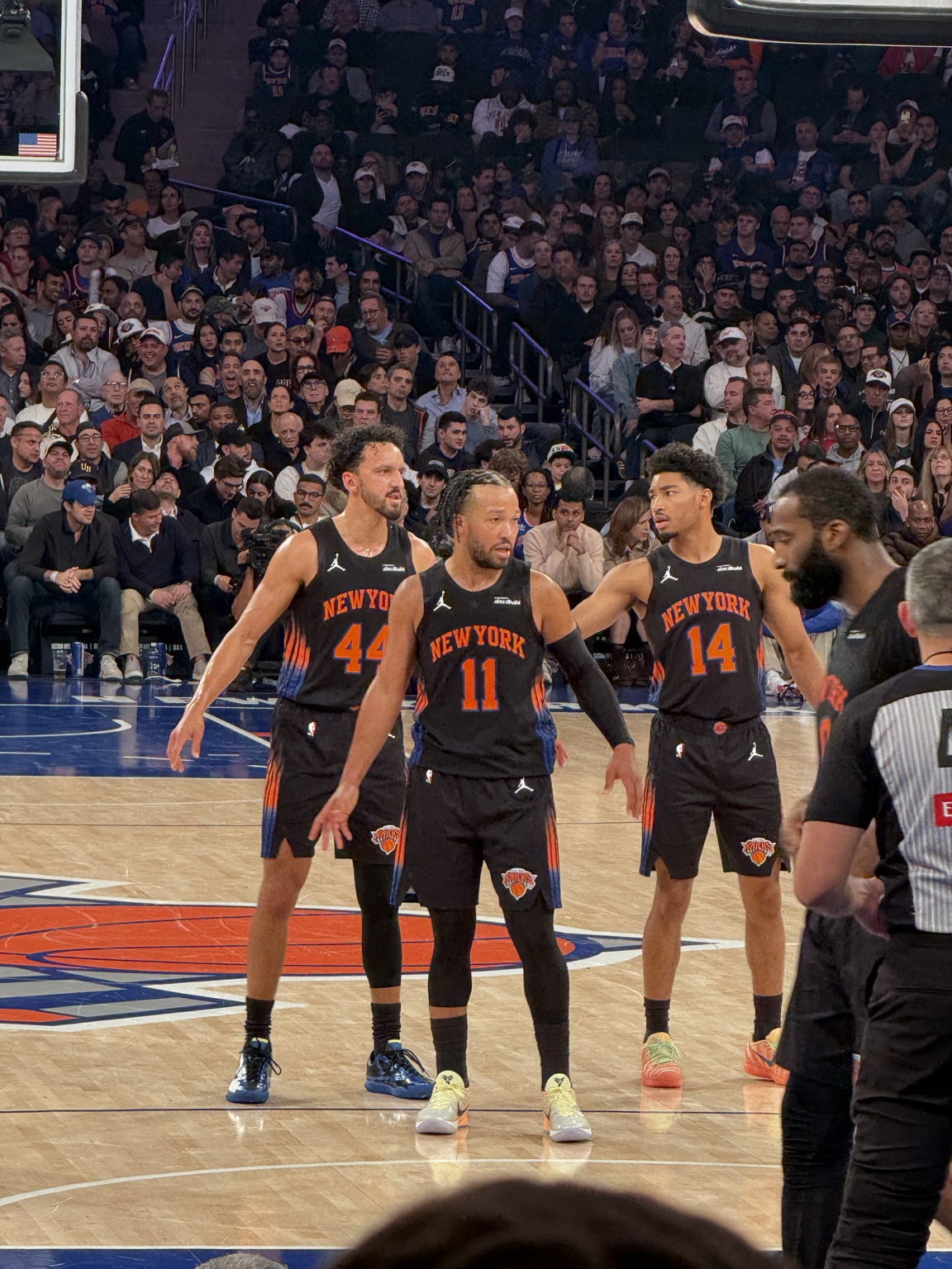 Knicks players on court in black New York uniforms, with spectators seated behind them.