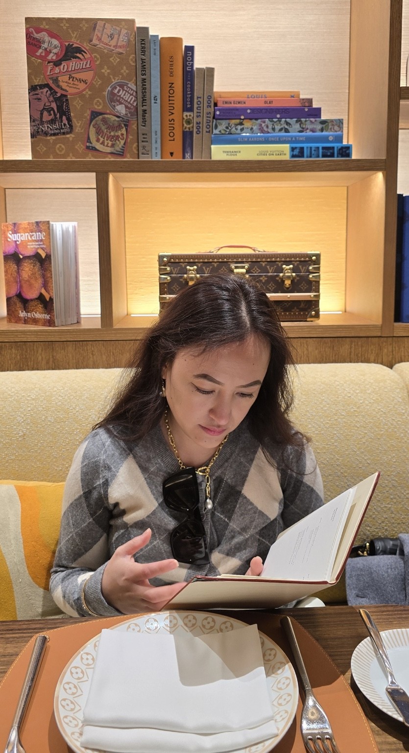 Frances browsing the menu at Le Café Louis Vuitton, with books and a monogrammed trunk on the shelves behind her.